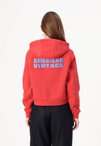 Red cropped hoodie made of textured fabric, featuring a large blue “AMERICAN VINTAGE” logo on the back. Hoodie has a drawstring.
