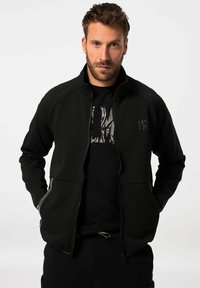 Schwarze Zip-up-Jacke mit hohem Kragen, seitlichen Taschen, glatter Textur und einem Logodetail auf der linken Brust. Getragen über einem schwarzen T-Shirt.