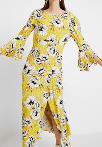 Abito maxi floreale giallo con maniche a campana, caratterizzato da motivi floreali in bianco e nero, un collo rotondo e uno spacco alto fino alla coscia.