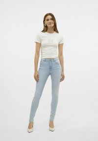 Camiseta blanca con diseño de mariposa, vaqueros ajustados de color azul claro y tacones beiges. La modelo está de pie con las manos en las caderas frente a un fondo liso.