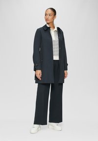 Námornícky trench coat s gombíkmi, špicatým golierom a dlhými rukávmi. V kombinácii s pruhovaným svetrom a voľnými nohavicami. Biele tenisky dokončujú celý outfit.