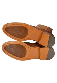Semelles de chaussures en cuir brun avec un motif embossé détaillé, bouts ronds et talons beige contrastants ornés de rivets métalliques.