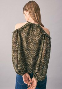Kvinde med langt brunt hår iført en off-shoulder, løs leopardprint-bluse og blå jeans, med hænderne samlet bag ryggen.