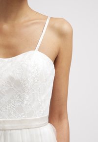 Abito bianco con corpino in pizzo, spalline sottili e cintura in satin. Presenta un sovrapposto semi-trasparente e un motivo floreale in pizzo sulla parte superiore.