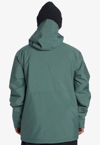 Vodotesný zelený kabát s voľným strihom, nastaviteľnou kapucňou a elastickými manžetami. Má hladkú textúru a minimalistický dizajn.