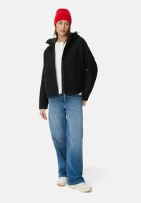 Zwarte zip-up jas met een capuchon, gecombineerd met een witte shirt en blauwe jeans. Model draagt een rode gebreide beanie en witte sneakers.