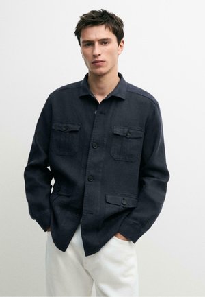 Jeune homme porte une veste bleu marine foncé à boutons avec quatre poches avant et un pantalon blanc, debout devant un fond clair uni.