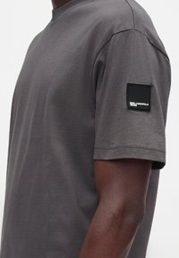 T-shirt gris foncé à manches courtes sur une personne montrant une étiquette noire avec un texte blanc "Karl Lagerfeld Jeans" sur la manche supérieure.