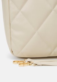 Gros plan sur le coin d'un sac en cuir matelassé beige avec deux boucles en métal doré attachées à des sangles assorties, posé sur une surface blanche.