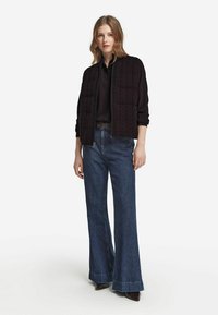 Kvinna klädd i svart stickad zip-up väst över en svart skjorta, tillsammans med hög midja vidbrutna blå jeans och mörka spetsiga skor.