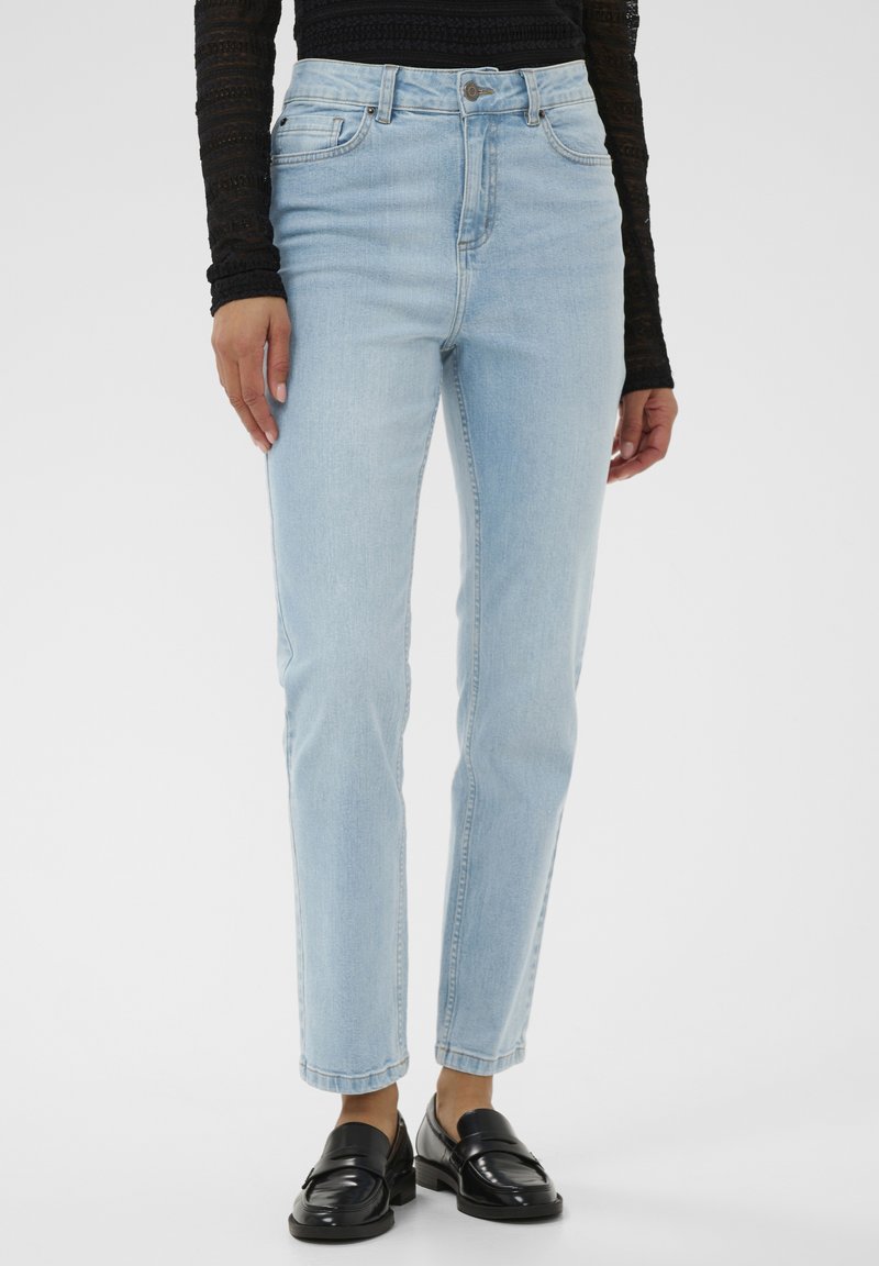 Femme portant un jean droit bleu clair, un haut en dentelle à manches longues noir et des mocassins en cuir noir, debout devant un fond blanc.