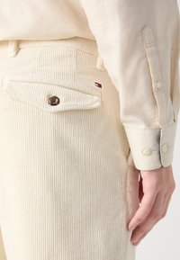 Beżowe spodnie z korduroyu z tylną kieszenią, z okrągłym guzikiem i małym logo w granatowym i czerwonym kolorze. W zestawie z jasnym, zapinanym na guziki koszulem.