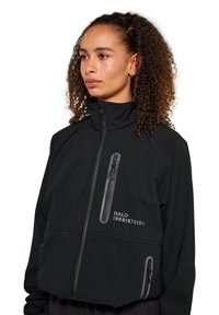 Svart softshell-jacka med hög krage, flera fickor med dragkedja och tryckt text. Har en slät textur och en modern design.
