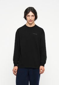 Černé tričko s dlouhým rukávem vyrobené z bavlny. Na hrudi se nachází logo značky a na levém rukávu text. Jednoduchý design s kulatým výstřihem.