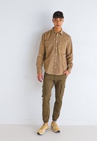 Camisa de botonada de pana color marrón claro con dos bolsillos en el pecho, combinada con pantalones cargo de color verde oliva y zapatillas beige. Gorra negra en la cabeza.