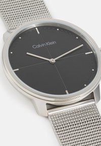 Reloj Calvin Klein con esfera negra, índices plateados y manecillas delgadas. Cuenta con una brillante correa de malla de acero inoxidable y un bisel pulido.