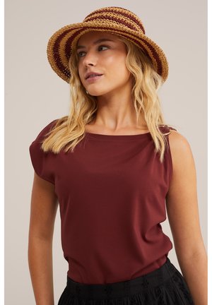 Naine, kes kannab bordoopunast varrukateta toppi ning triibulist õlgkübarat, vaadates küljele, lahtiste blondide juustega üle õlgade.