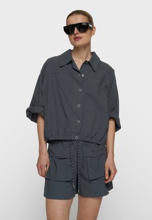 Mujer con camisa de botones gris oscuro de gran tamaño con mangas remangadas, pantalones cortos a juego con grandes bolsillos y gafas de sol negras de gran tamaño.