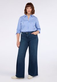 Camicia button-up azzurro chiaro con ricamo di piccoli cuori rossi, abbinata a jeans dark denim a vita alta e gamba larga e scarpe da ginnastica color crema.