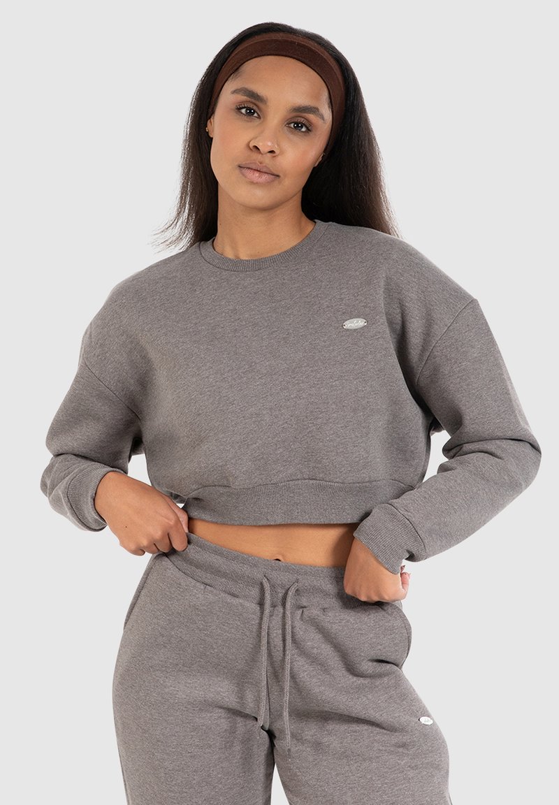Vrouw met lang haar, een bruine haarband, een grijze cropped sweatshirt en bijpassende grijze sweatpants voor een effen lichte achtergrond.