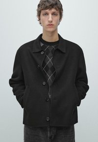 Sort jakke i uldblanding med krave, knaplukning foran og sidelommer. Båret over en grå sweater med argylemønster.