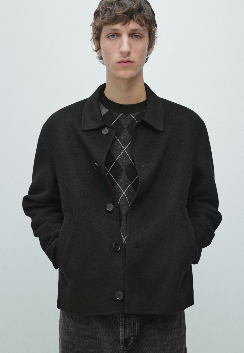 Sort jakke i uldblanding med krave, knaplukning foran og sidelommer. Båret over en grå sweater med argylemønster.