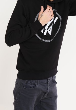 Personne portant un sweat à capuche noir avec un graphisme blanc et un jean foncé, main touchant la poitrine montrant des tatouages sur les doigts et le poignet.