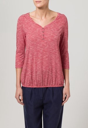 Czerwona koszula Melange z długim rękawem, dekoltem w serek, trzema guzikami, marszczonymi ramionami i elastycznym dołem, zestawiona z granatowymi spodniami.