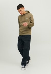 Felpa verde oliva con tasca frontale, coulisse regolabile e dettaglio del logo, abbinata a pantaloni cargo scuri e sneakers nere.