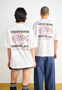 White oversized t-shirt featuring "VERTERE" and "BERLIN" text, with colorful graphic designs on the back; worn with striped socks and jeans.