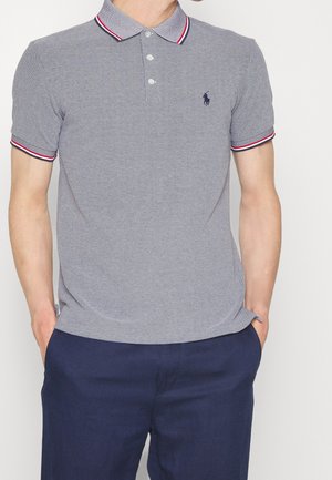 Man die een grijs poloshirt met korte mouwen met rode en witte biezen en donkerblauwe broek draagt, handen in de zakken, staand tegen een lichte achtergrond.