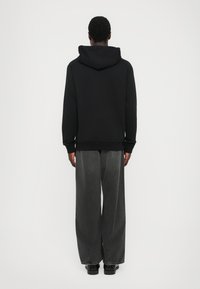 Persona con una sudadera negra y jeans oscuros holgados, de pie de espaldas frente a un fondo blanco liso.