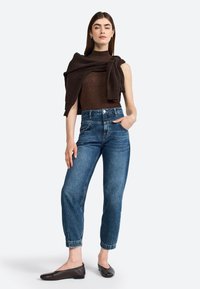 Dark brown sleeveless top with a knitted sweater draped around the shoulders, paired with light blue high-waisted jeans and brown flats.