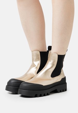 Botines metálicos dorados con un panel lateral elástico negro, suela de goma negra gruesa y un detallado texturizado en la suela.