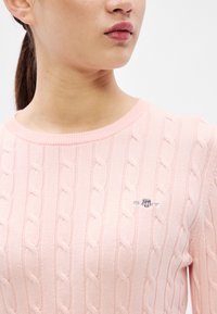 Ružový sveter s vrkočovým vzorom, rebrovaným výstrihom a vyšitým logom na hrudi. Má jemnú textúru a priliehavý strih.