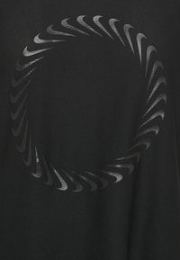 Musta urheilupaita, jossa on ympyrämuotoinen harmaista viuhkoista koostuva kuvio rinnan keskellä ja teksturoitu kangaspinta.