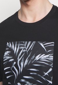 Primer plano de una persona que lleva una camiseta negra con un estampado monocromo de hojas tropicales en el frente.