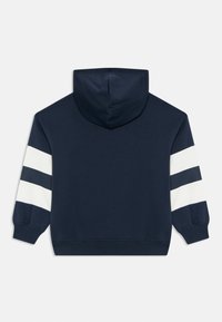 Felpa blu navy con strisce bianche e blu navy sulle maniche, polsini a coste e cappuccio, mostrata da dietro su uno sfondo bianco.