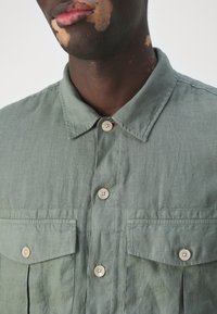 Grünes Hemd aus leichtem Stoff mit spitzem Kragen, zwei Vorderentaschen und abgerundeten Knöpfen. Weiche Textur.