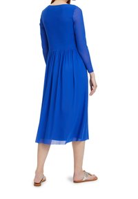 Långärmad blå klänning med rund halsringning, flödande kjol och genomskinliga ärmar, bärs med platta sandaler. Mjuk, lätt tyg.