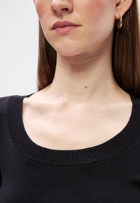 Γυναίκα με μ�αύρο πουλόβερ με στρογγυλή λαιμόκοψη, ίσια καστανά μαλλιά, χρυσά σκουλαρίκια κρίκοι και φυσικό μακιγιάζ που δίνει έμφαση στα χείλη και τον λαιμό.