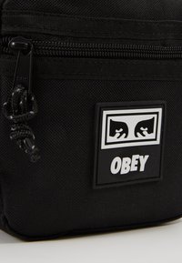 Czarna torba z materiału o fakturowanej powierzchni, z górnym zamkiem błyskawicznym oraz prostokątną gumową naszywką z białym napisem "OBEY" i stylizowanymi oczami.
