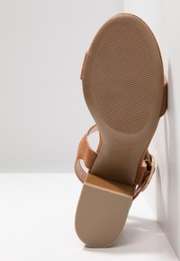 Sandale en daim marron inclinée contre un mur blanc montrant la semelle texturée avec le logo "NEW LOOK" et les détails de taille 4/37.