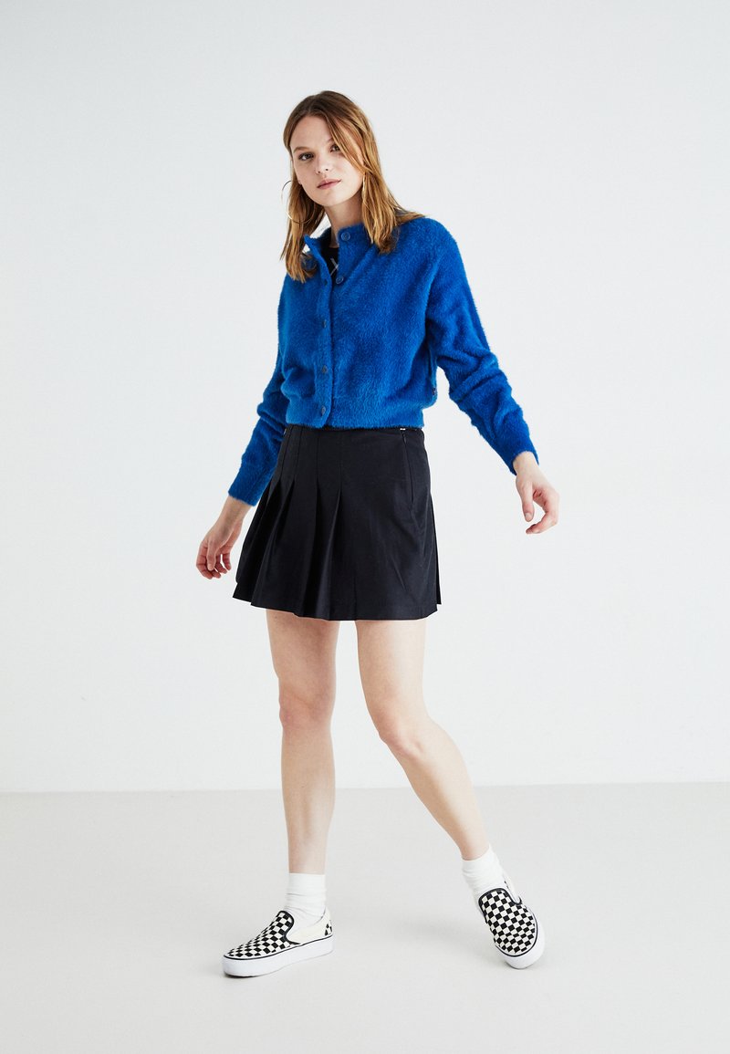 Cardigan blu fluffy, gonna nera a pieghe e scarpe slip-on bianco e nere a scacchi. Il modello si trova su uno sfondo bianco.