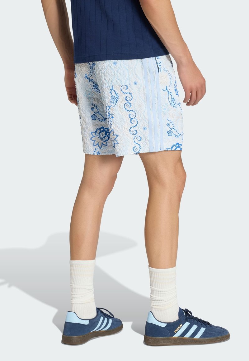 Man draagt textuur lichtblauwe korte broek met een bloemenpatroon, marineblauwe sneakers met lichtblauwe strepen, witte sokken en een marineblauw shirt.