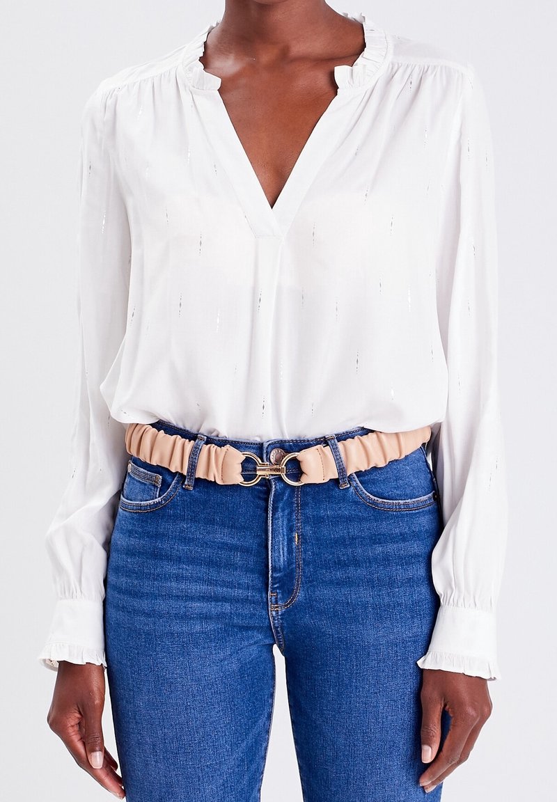 Femme portant un chemisier blanc à manches longues rentré dans un jean bleu avec une ceinture beige froncée dotée d'une boucle métallique dorée.