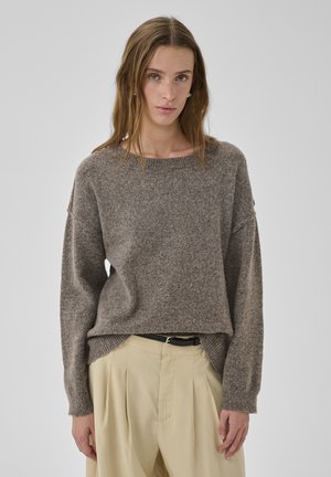 Mujer joven vestida con un suéter gris suelto y pantalones plisados beige, de pie contra un fondo gris sencillo.
