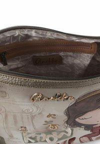 Strukturierte beiger Handtasche mit goldenen Akzenten. Das Innere zeigt ein Blumenmuster, eine Reißverschlusstasche und ein Etikett mit der Aufschrift "Anekke".