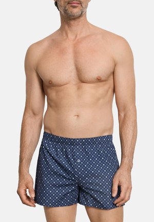 Hombre sin camisa que lleva puestos unos bóxers azules con un patrón y una cintura elástica, posando contra un fondo blanco liso.