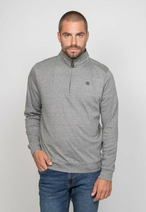 Homme aux cheveux courts et à la barbe, porte un pull gris à fermeture quart-zip et un jean bleu, debout avec une main dans la poche devant un fond uni.